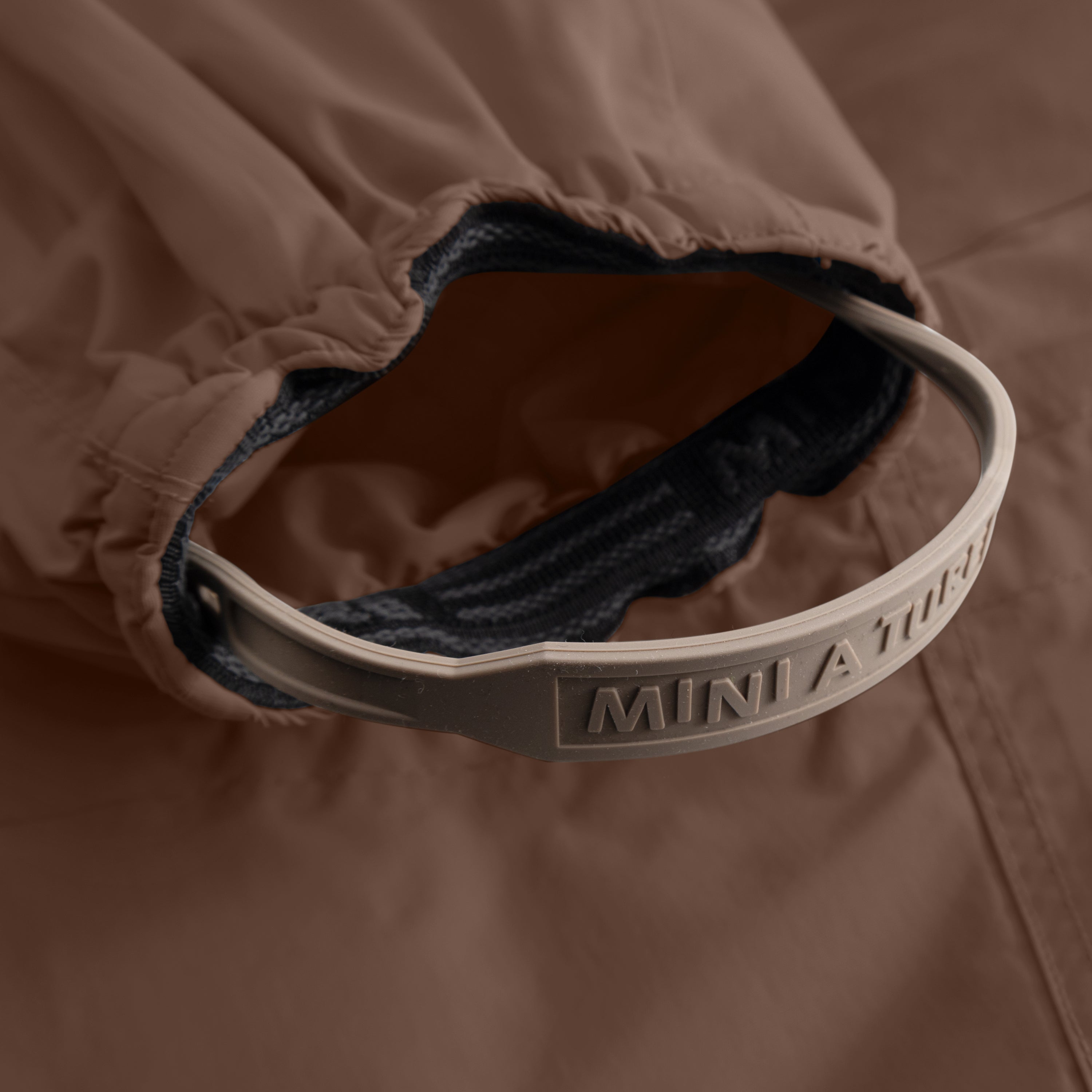 Detail of MATWISTI fleece lined snowsuit fur in brown showing the adjustable and detachable foot strap with embossed MINI A TURE branding on the taupe silicone band, elasticated ankle cuff with dark fleece lining visible inside, and beige outer fabric.