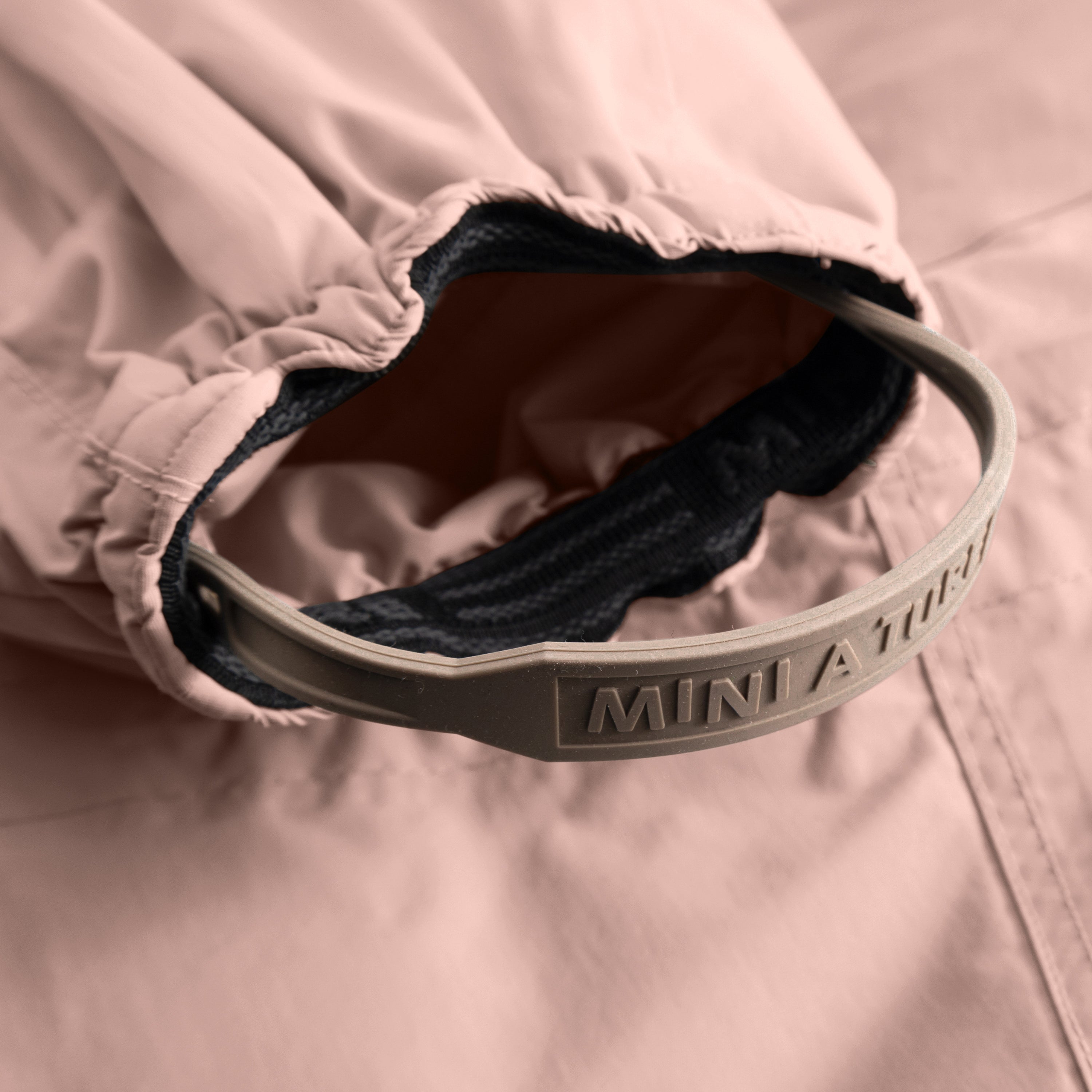 Detail of MATWISTI fleece lined snowsuit fur in soft pink showing the adjustable and detachable foot strap with embossed MINI A TURE branding on the taupe silicone band, elasticated ankle cuff with dark fleece lining, and beige fleece interior visible against the waterproof outer shell.