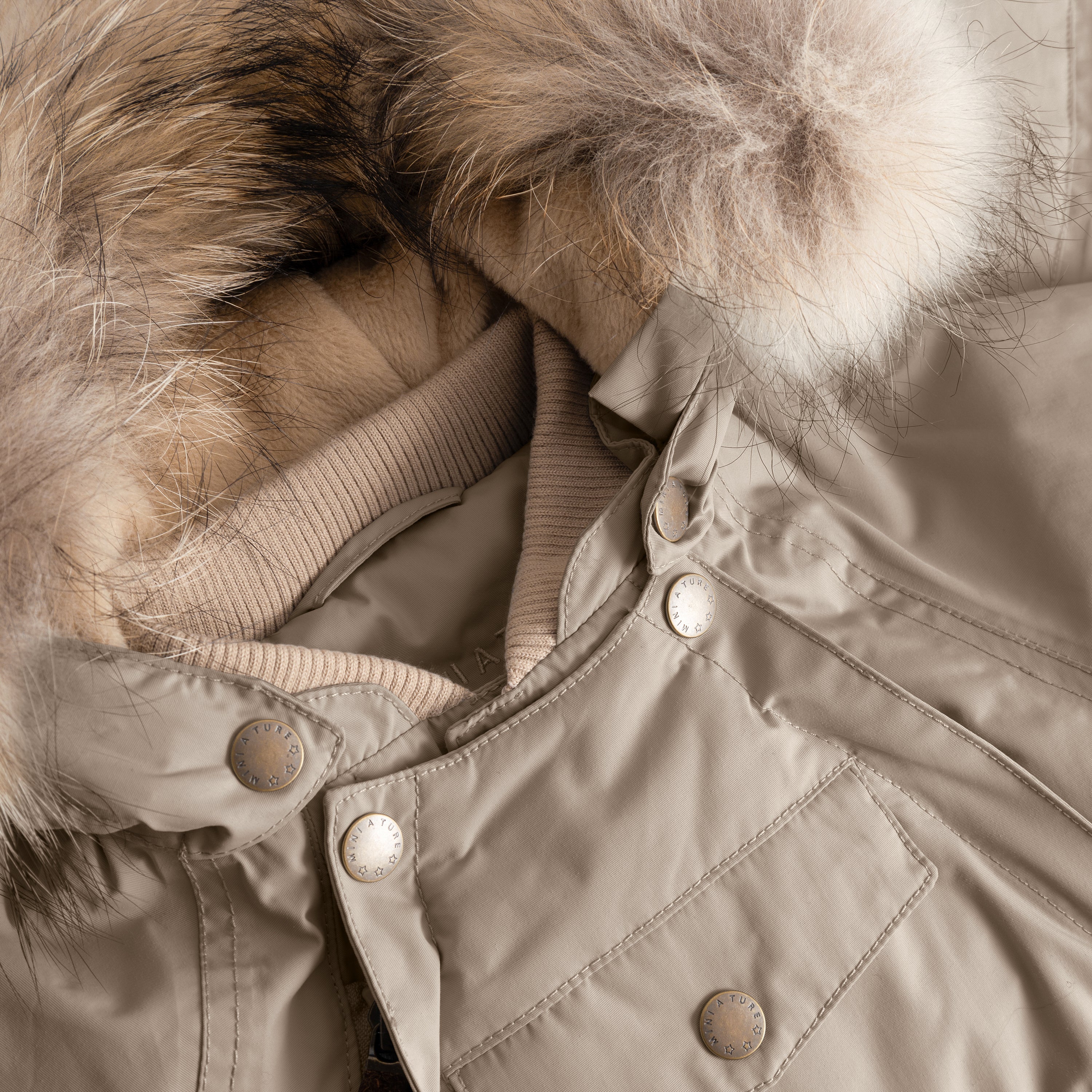 Close-up detail of MATWISTI fleece lined snowsuit fur showing the detachable genuine fur hood trim, ribbed collar, press-stud fastenings with MINI A TURE branding, and the taupe waterproof outer shell with chest pocket.