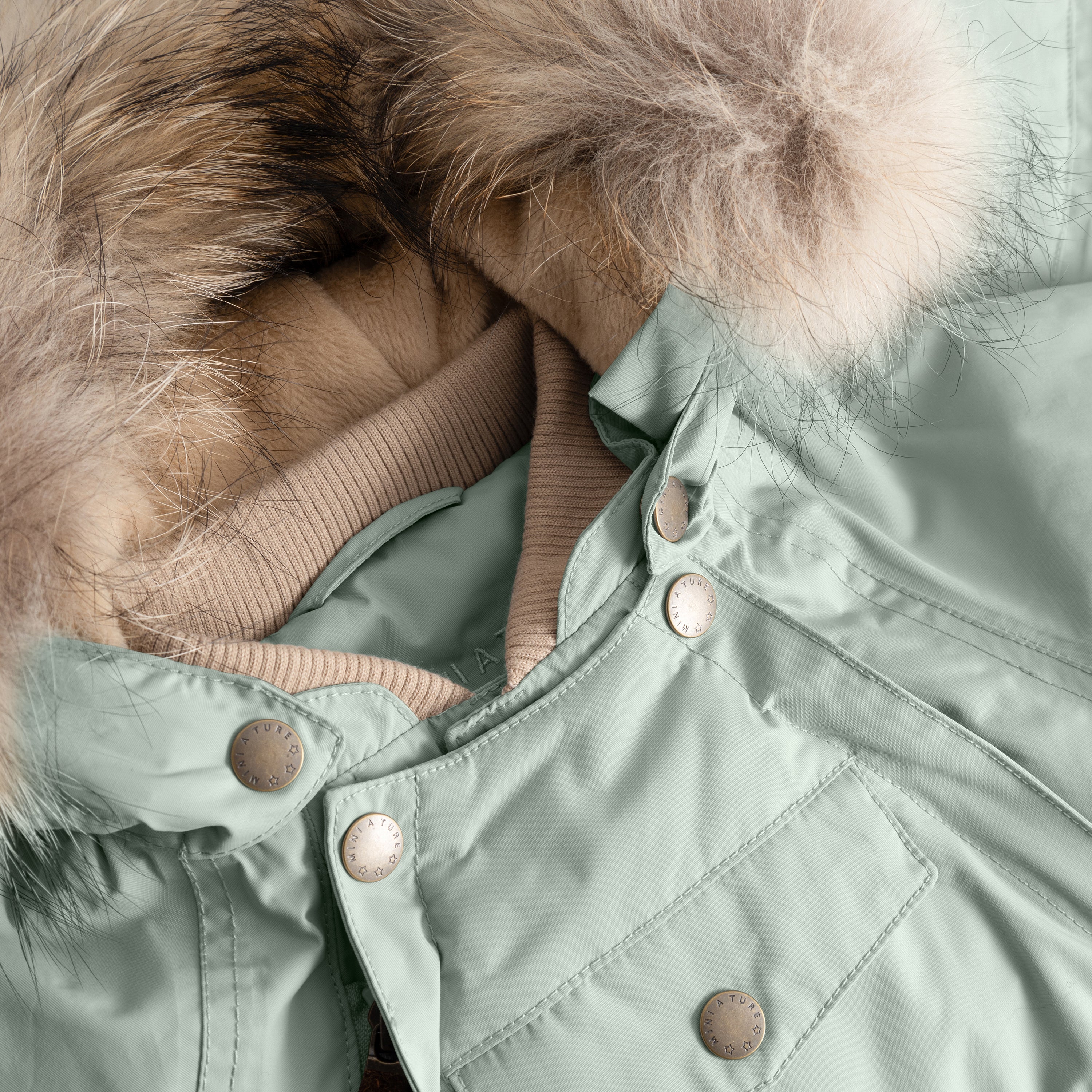 Close-up of MATWISTI fleece lined snowsuit fur in sage green showing the detachable genuine fur hood trim, taupe ribbed collar, MINI A TURE branded press-stud fastenings along the chest, and front pocket detail on the waterproof shell.