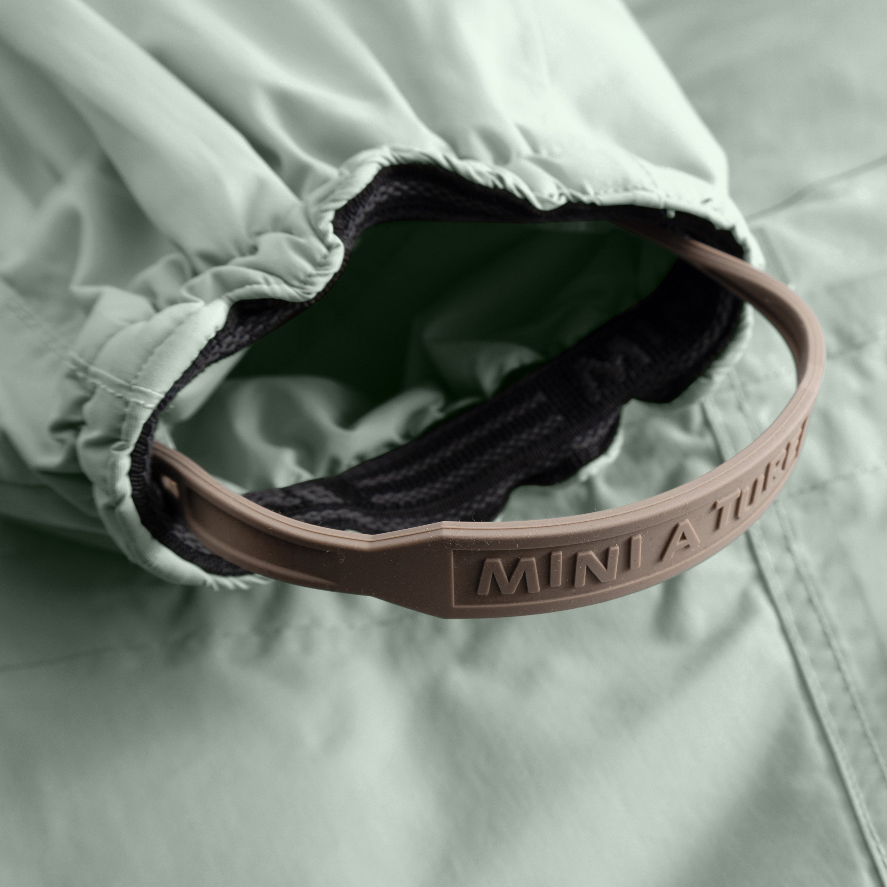 Detail of MATWISTI fleece lined snowsuit fur in sage green showing the adjustable and detachable foot strap with embossed MINI A TURE branding on the taupe silicone band, elasticated ankle cuff with dark fleece lining, and beige fleece interior visible against the waterproof outer shell.