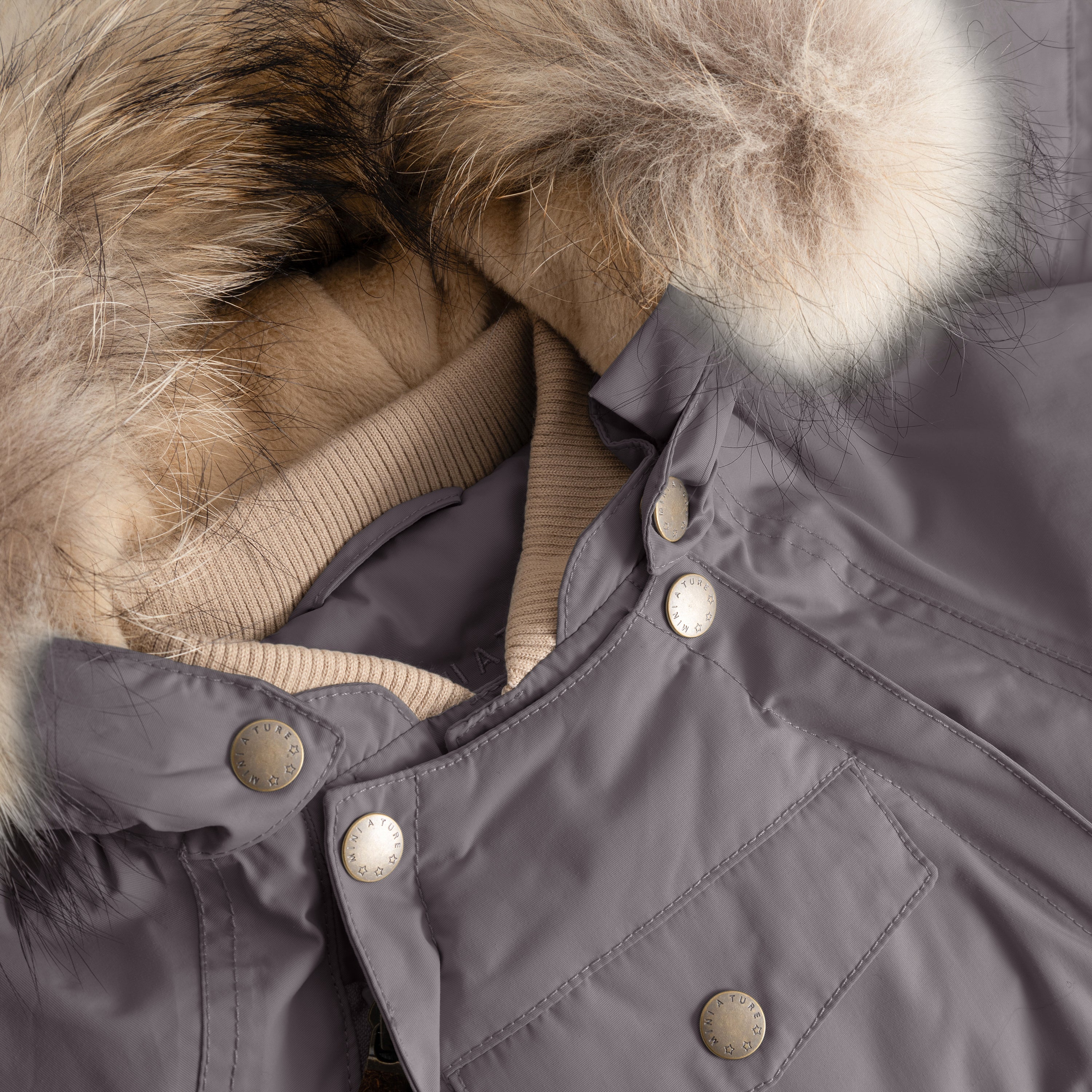 Detail of MATWISTI fleece lined snowsuit fur in grey showing the genuine fur hood trim, beige ribbed collar, MINI A TURE branded press-stud fastenings along the chest, and front pocket detail on the waterproof shell.