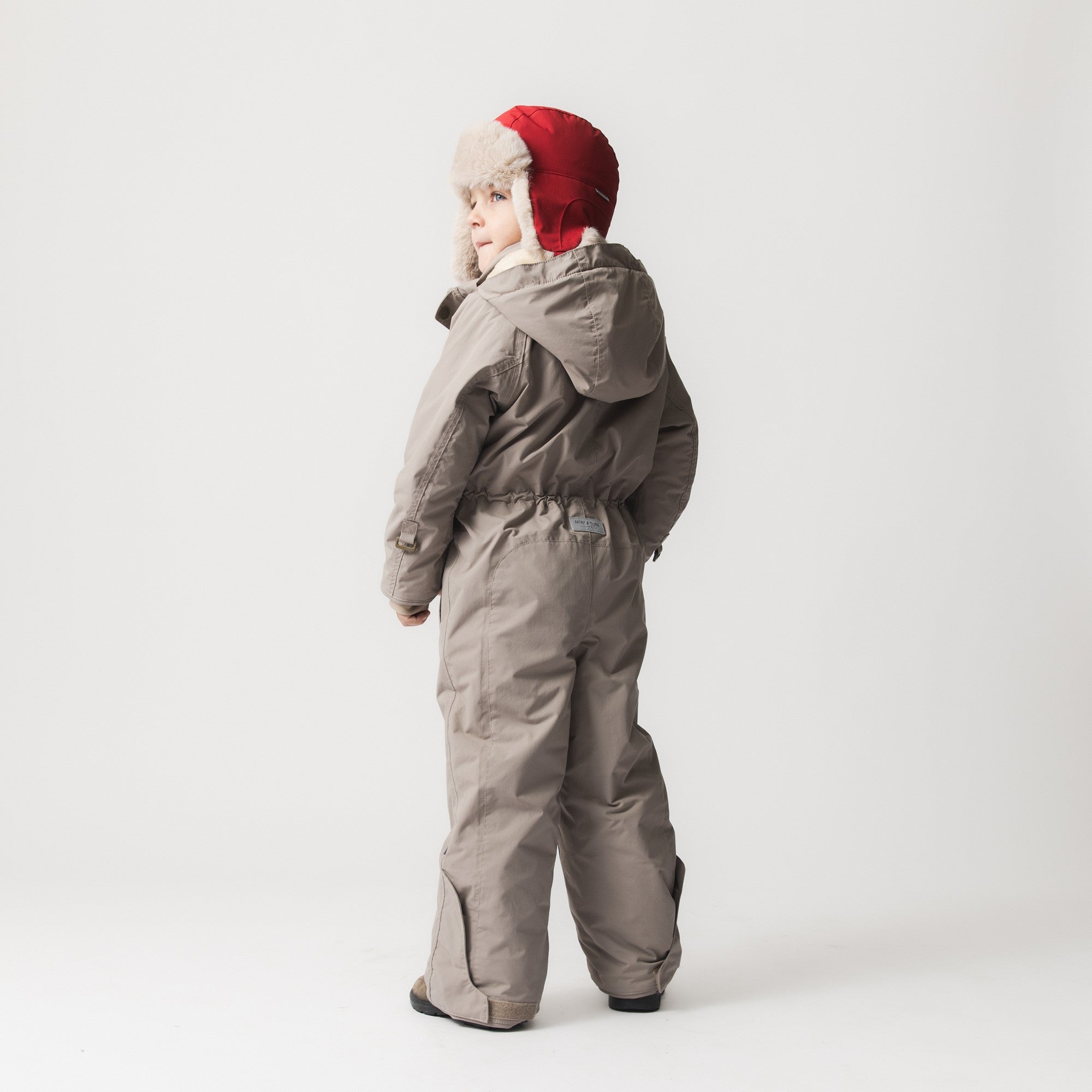 MATWANNI fleece lined snowsuit in grey worn by a child with a red faux-fur trimmed hat, photographed from behind to show the reinforced back panel, internal suspenders at the shoulders and removable foot straps at the ankles.