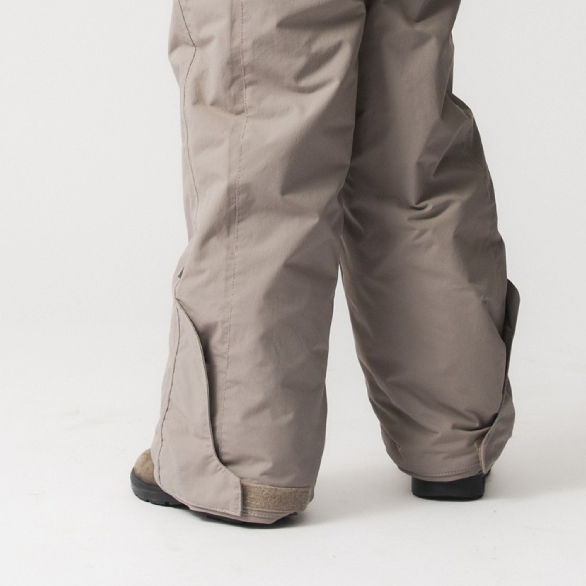 MATWANNI fleece lined snowsuit in beige shown from behind at ankle level, displaying the removable foot straps positioned over boots, reinforced lower leg construction and the waterproof recycled polyamide shell with fully taped seams.