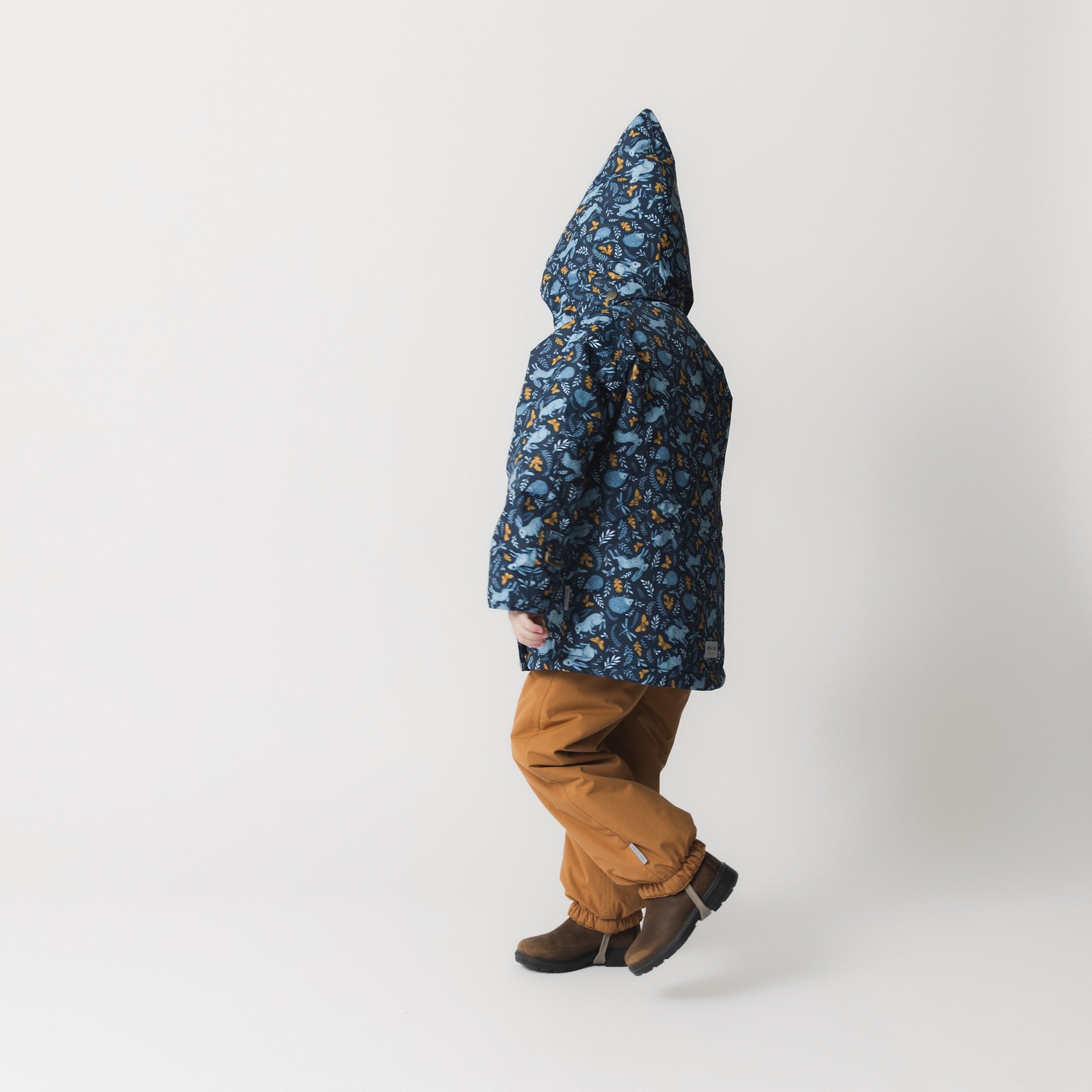 MATWANG fleece lined winter jacket shown in profile on a child with the hood up, revealing the jacket's silhouette and the Scandinavian floral print with blue rabbits and golden oak leaves, paired with mustard trousers and brown boots.