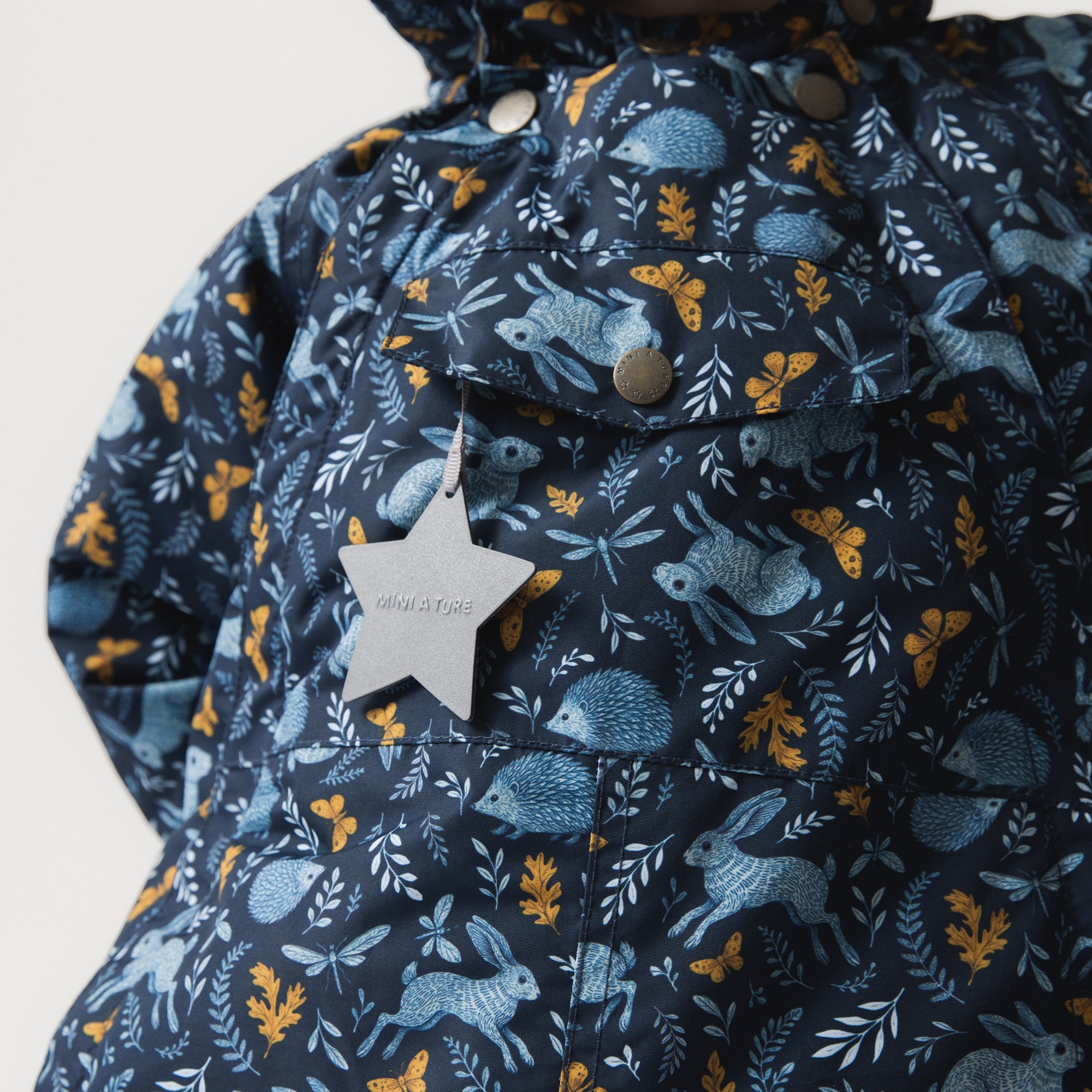 MATWANG fleece lined winter jacket photographed in close detail showing the front chest pocket with a press-stud fastening and the white reflective star hanging from a loop, set against the Scandinavian floral print featuring blue rabbits, golden oak leaves and botanical details across the waterproof shell.