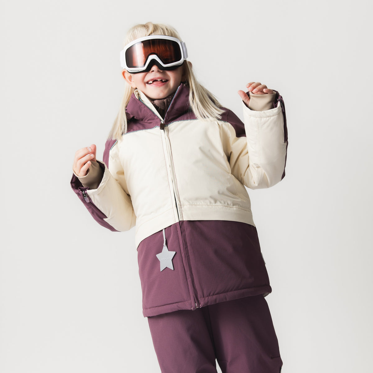 MATADANE ski tech jacket worn by a child with ski goggles, shown in plum and cream colourblock styling with a front zip, chest pocket featuring a reflective star, and matching ski trousers.