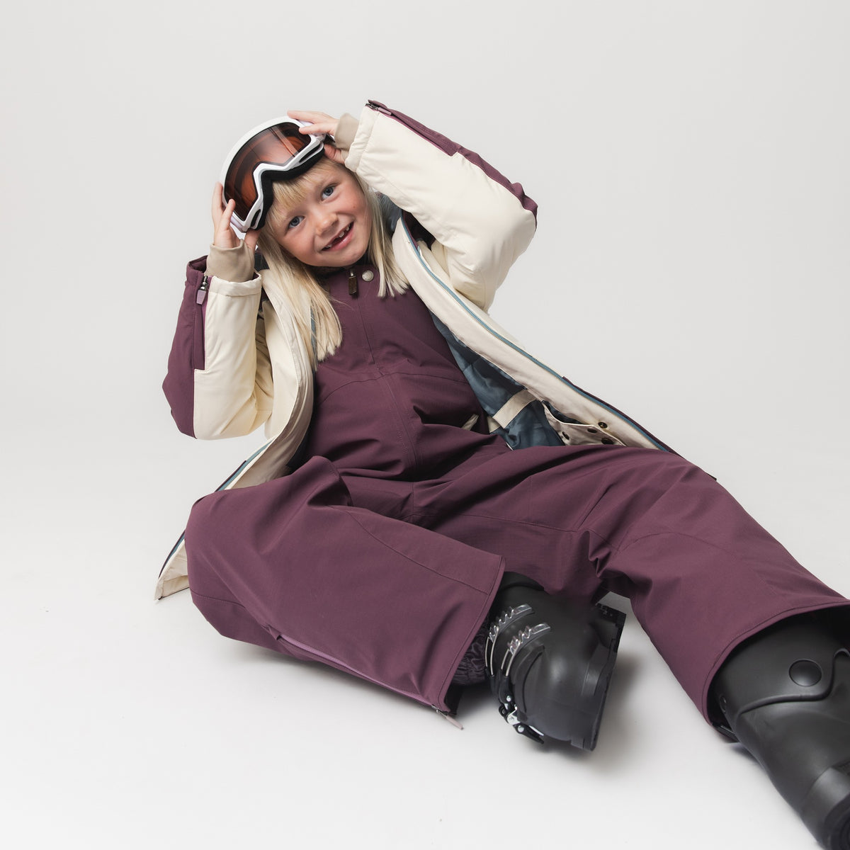 MATADANE ski tech jacket worn by a child seated on the floor with ski goggles and matching burgundy ski trousers, showing the jacket fully unzipped to reveal the internal powder skirt and insulated lining in cream and teal colourblock styling.