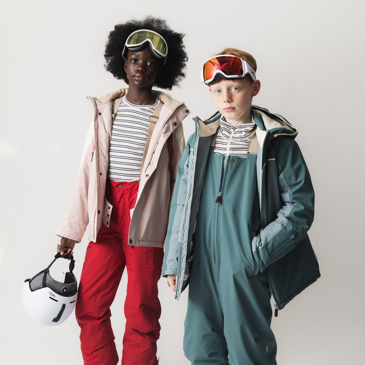 MATADANE ski tech jacket in pale pink worn by a child with ski goggles and red ski trousers, shown alongside another child in teal ski jacket, both holding ski helmets and wearing striped base layers.