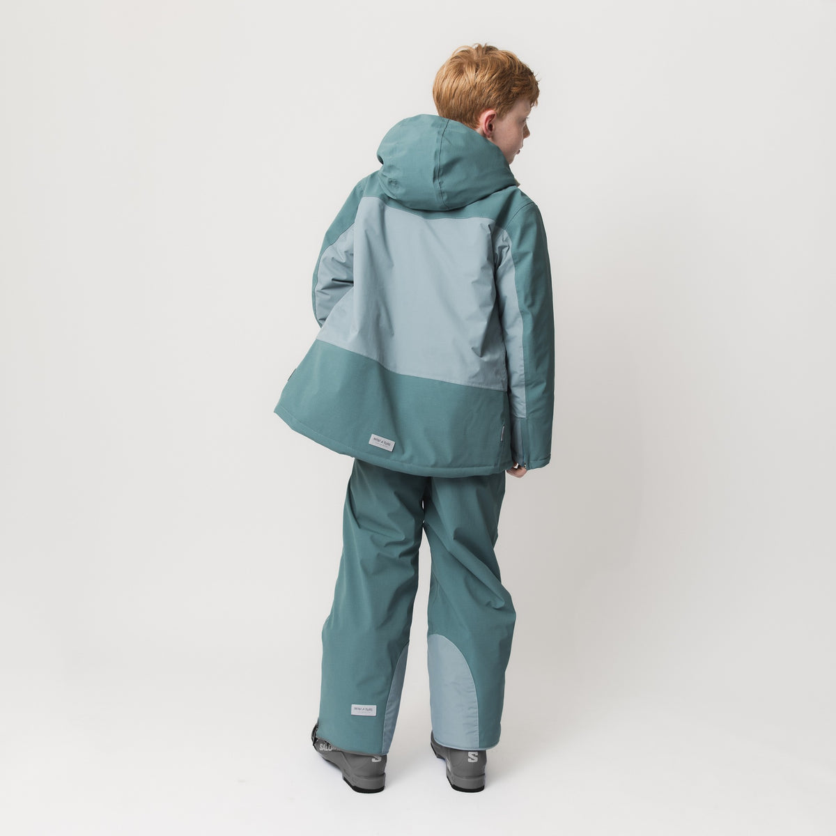 Kind in blaugrüner MATADAL-Skihose mit hellblauen Verstärkungen am Knie und passender Jacke mit Kapuze, Rückansicht, zeigt die wasserdichte Ripstop-Konstruktion und THERMOLITE-Isolierung in Skischuhen.