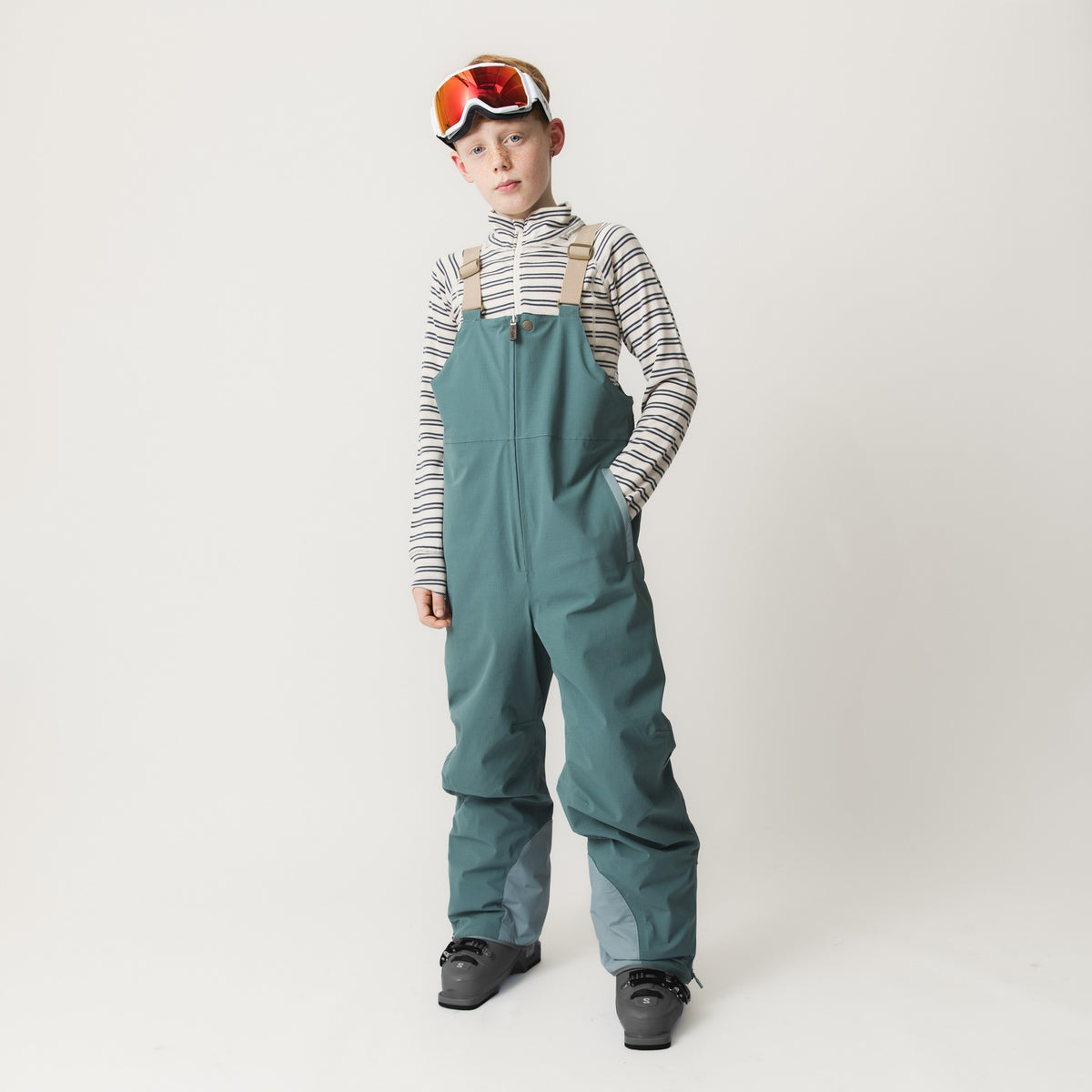 Kind in blaugrüner MATADAL-Skihose mit beigen Trägern und gestreiftem Pullover, trägt Skibrille und Skischuhe, zeigt die wasserdichte Ripstop-Konstruktion und THERMOLITE-Isolierung.