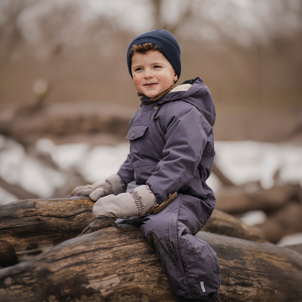 Young child wearing the MATWISTI fleece lined snowsuit in charcoal grey with a navy knitted beanie and cream mittens, sitting on driftwood outdoors, showing the one-piece construction, front chest pocket and elasticated leg openings with adjustable foot straps.