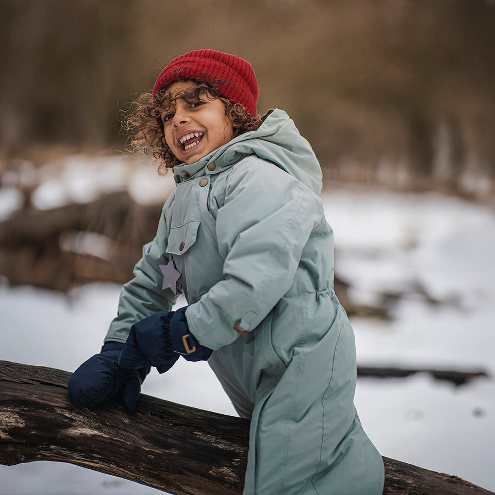 Child laughing whilst climbing on a fallen log in a snowy woodland setting, wearing the MATWISTI fleece lined snowsuit in sage green with a red knitted beanie and navy mittens.