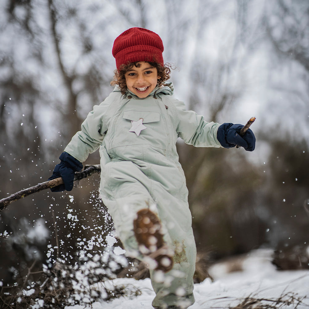 Child playing outdoors in snowy woodland wearing the MATWISTI fleece lined snowsuit in pale blue with a red knitted beanie and navy mittens, holding sticks whilst kicking up snow.