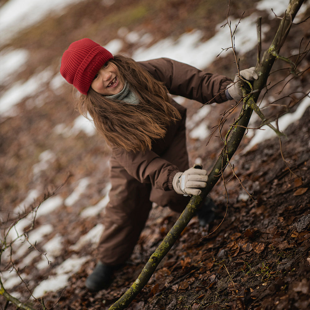 MATWANNI fleece lined snowsuit in brown worn by a child climbing a moss-covered branch outdoors, paired with a red knitted beanie, grey neck warmer and light-coloured gloves, captured from above during active winter play.