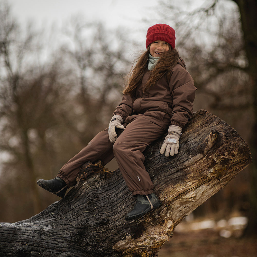 MATWANNI fleece lined snowsuit in brown worn by a child sitting on a fallen tree trunk outdoors, paired with a red knitted beanie, grey neck warmer and light-coloured gloves, with the removable foot straps visible over dark winter boots.