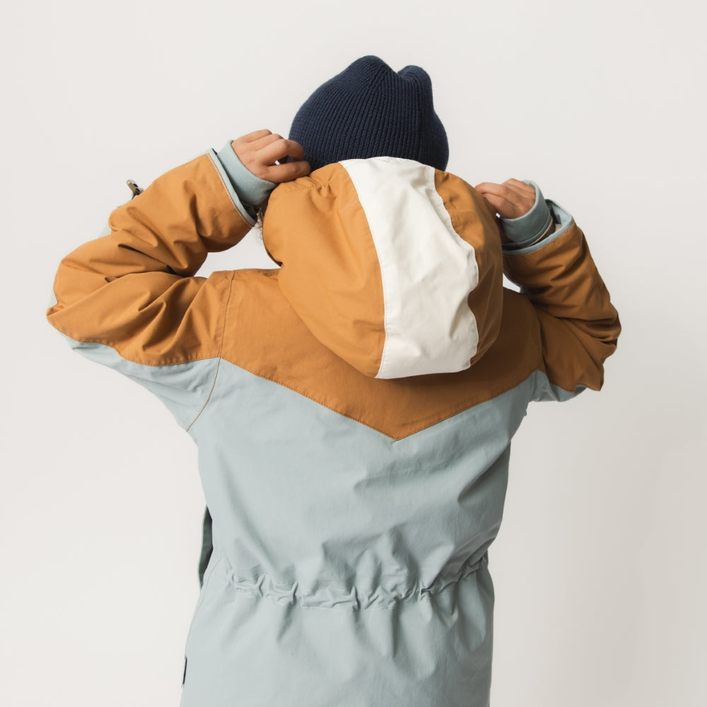Child pulling on the MATWALDO fleece lined winter jacket over their head, highlighting the hidden centre-front zip, adjustable internal waist and the practical colourblock design in brown sugar, baby blue and cream during everyday dressing.