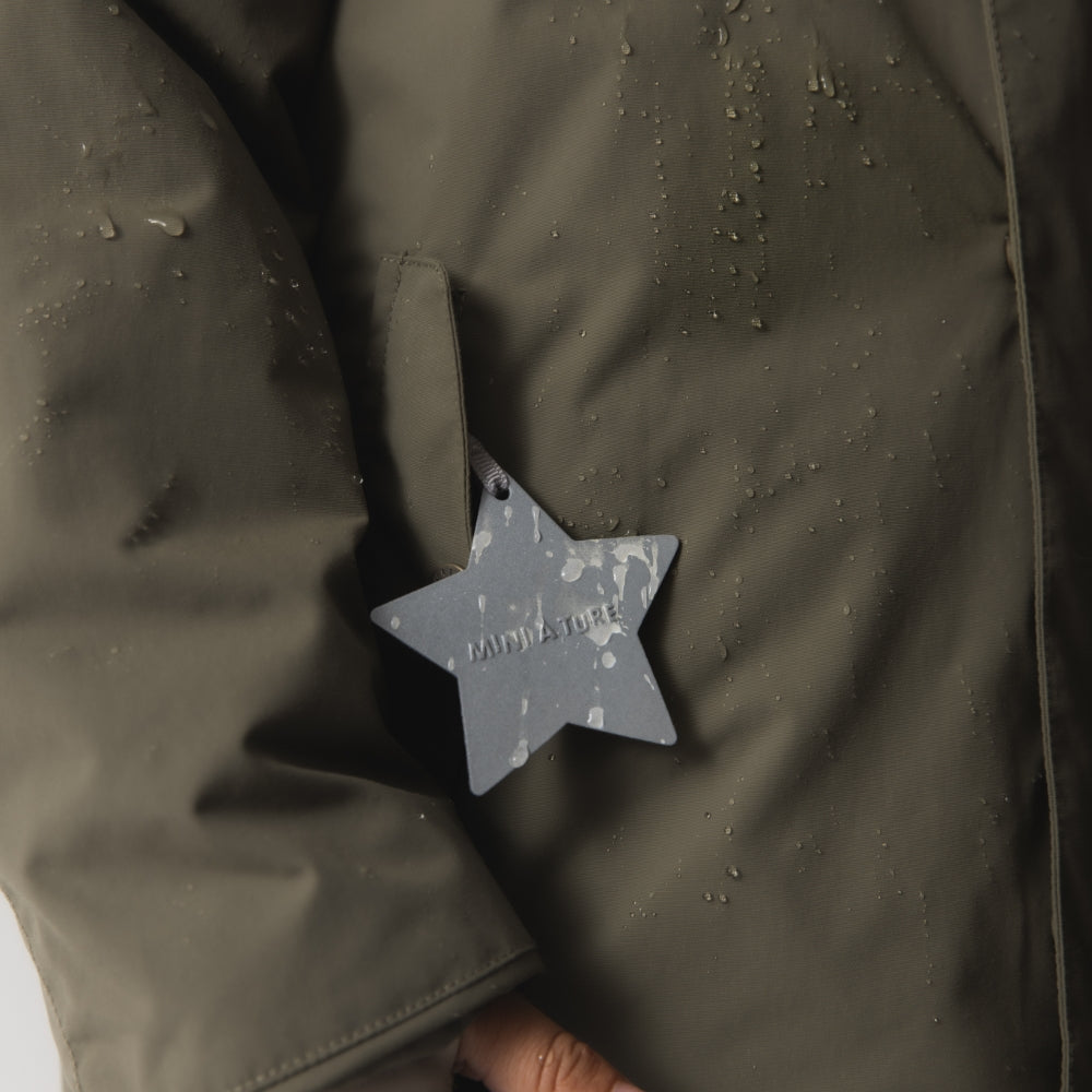 Close-up of the MATWALLY fleece lined winter jacket showing water droplets beading on the olive green recycled polyamide outer fabric, with the white reflective star detail embossed with MINI A TURE branding on the right pocket.