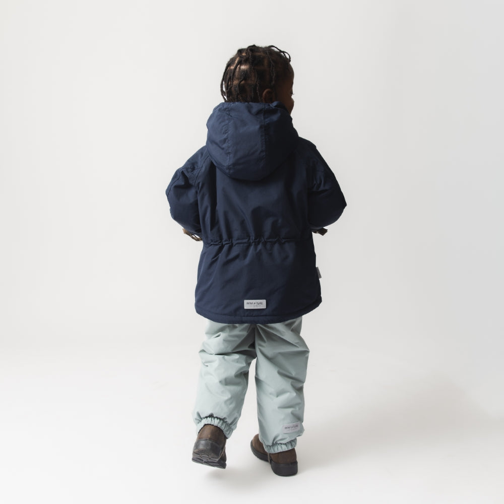 Rear view of a young child wearing the MATWALLY fleece lined winter jacket in navy blue, paired with light grey trousers, showing the detachable hood, adjustable internal waist and the white MINI A TURE woven label at the centre back hem.