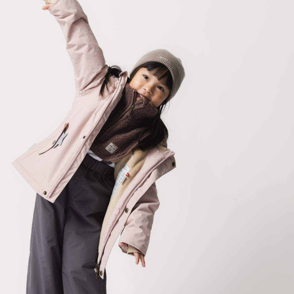 Young child wearing the MATWALLY fleece lined winter jacket in dusty pink over a brown fleece mid-layer and charcoal trousers, with one arm raised overhead in a playful pose, demonstrating the jacket's freedom of movement and layering capability.