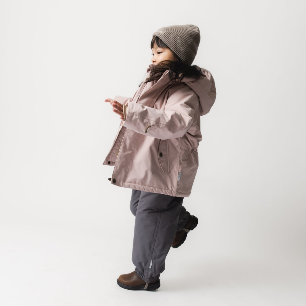 Young child wearing the MATWALLY fleece lined winter jacket in dusty pink with detachable hood, paired with charcoal trousers, a grey knitted beanie and brown boots, shown in profile with hands clasped together, demonstrating the jacket's regular fit and layering over everyday clothing.