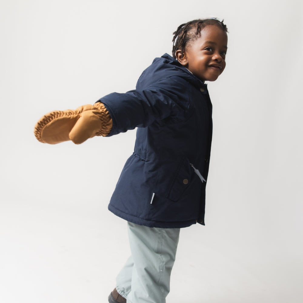 Young child wearing the MATWALLY fleece lined winter jacket in navy blue, photographed mid-movement with one leg raised, showing the jacket's regular fit and freedom of movement, paired with light grey trousers and tan boots.