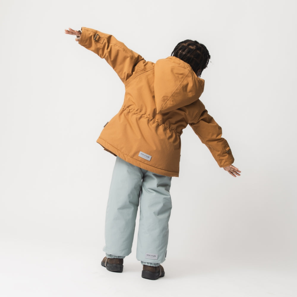 Child seen from behind wearing the MATWANG fleece-lined winter jacket in warm brown with arms outstretched, paired with light grey trousers and brown boots, showing the white MINI A TURE label at the hem and the jacket's regular fit during movement.