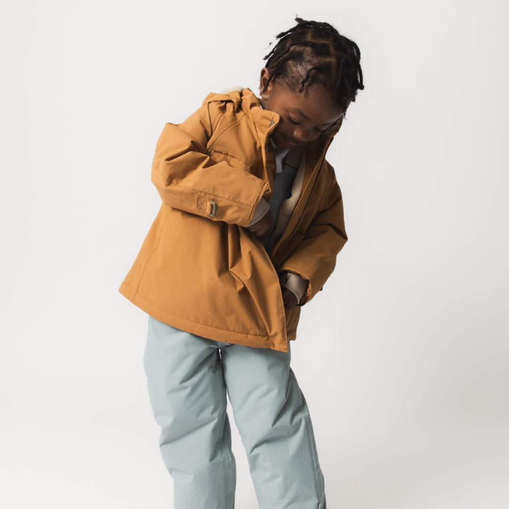 Child wearing the MATWANG fleece-lined winter jacket in warm brown with hood down, paired with pale blue trousers, demonstrating the jacket's regular fit and freedom of movement during active play.