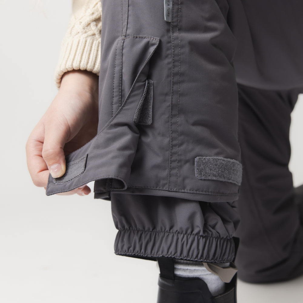 MATWITTE snow pants in charcoal grey showing the adjustable ankle opening with hook-and-loop fastening, elasticated cuff, integrated foot strap and reinforced lower leg construction designed to fit over ski boots.